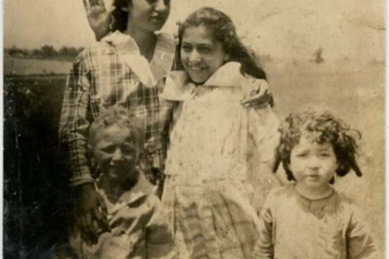 Four children pose outdoors in old-fashioned clothes, smiling and standing close together on a sunny day.