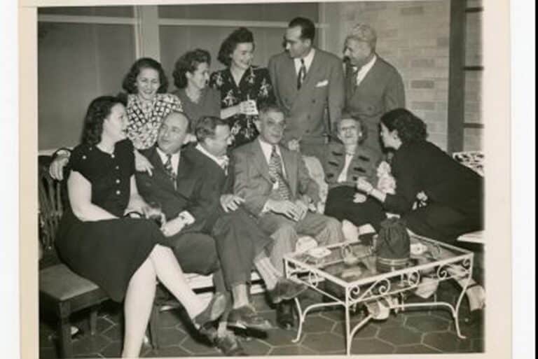 A group of men and women in formal attire gather, smiling and chatting in a living room setting.