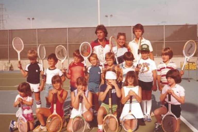 A group of kids and adults posing with tennis rackets on an outdoor court, smiling for the camera.