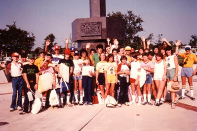 A large group of teens in casual ’80s outfits smiles and waves outdoors in front of a monument.