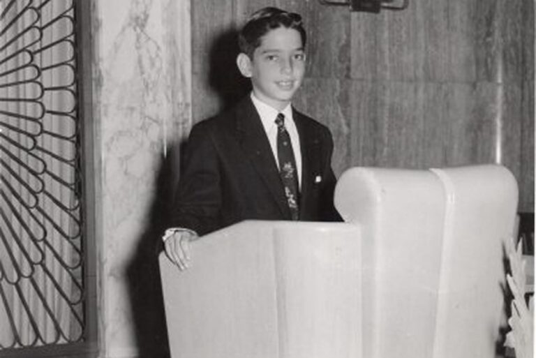 A boy in a suit stands at a podium in a formal setting, smiling for a black-and-white photo.