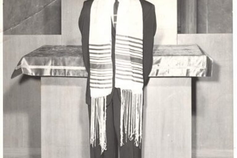 A person in a suit and tallit stands in front of an altar, facing the camera with hands behind their back.