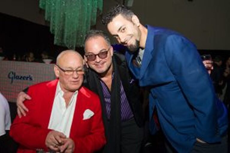 Three men wearing suits pose together and smile at an indoor event under green hanging decor.