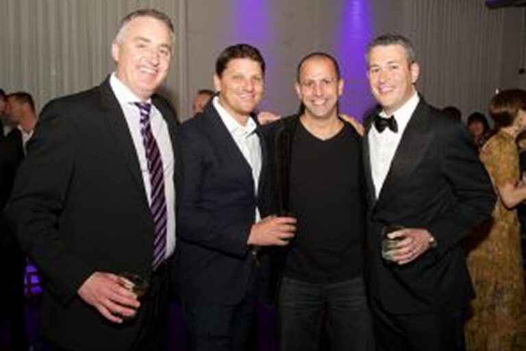 Four men in suits and tuxedos smile at a party, holding drinks, with purple lighting in the background.