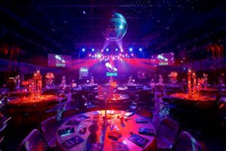 A banquet hall with round tables, dim lighting, a disco ball, and colorful stage lights overhead.