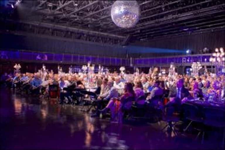 A large audience sits at tables under a disco ball in a dimly lit, purple-hued event hall.
