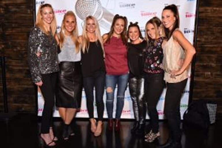 Seven women stand together smiling in front of a step-and-repeat backdrop at an indoor event.