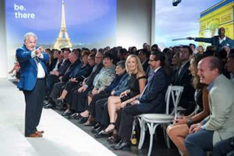 A man gestures on a runway in front of an audience; Eiffel Tower image and “be. there” text appear in the background.