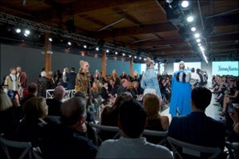 Models walk a runway at a fashion show, with a seated audience and bright overhead lights.