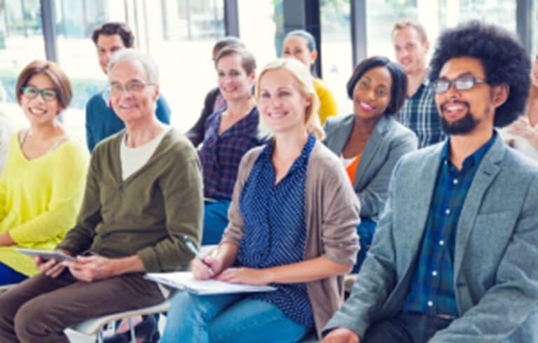 A diverse group of people sit in chairs, smiling and listening attentively in a bright room.