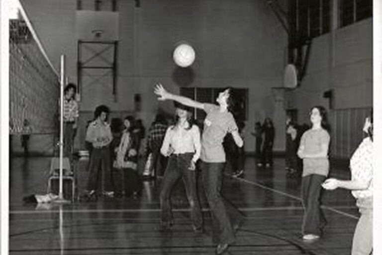 A group of people play volleyball in a gymnasium while others watch in the background.