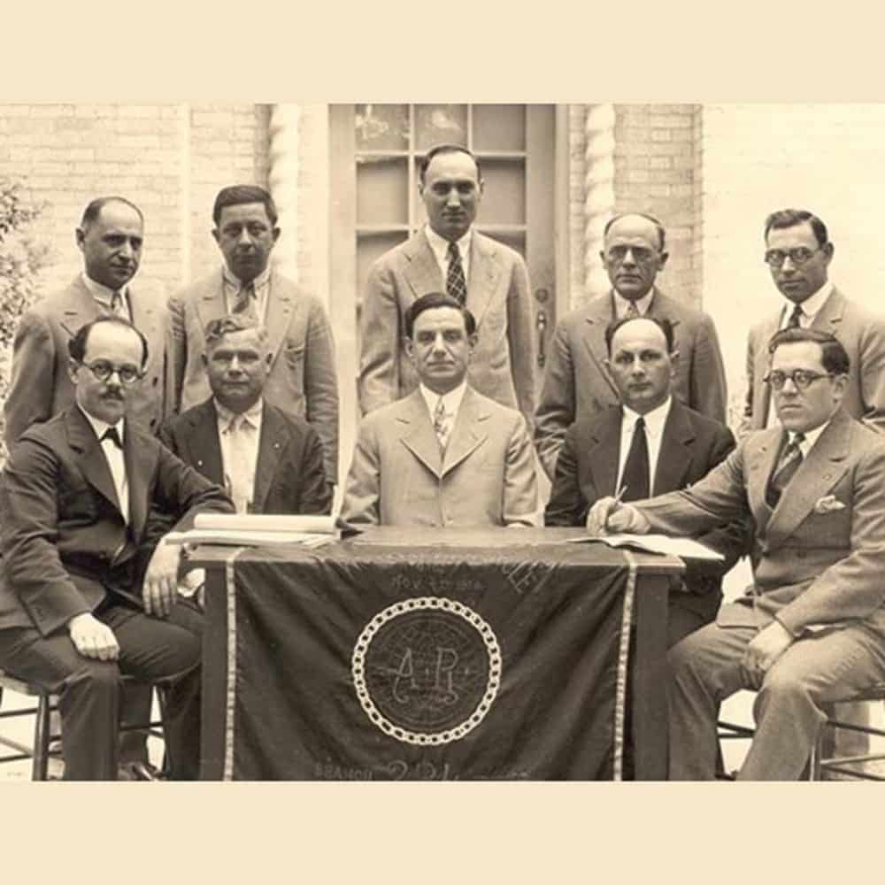 Ten men in suits pose around a table with a cloth featuring a circular emblem and “AP” letters, in front of a brick building.