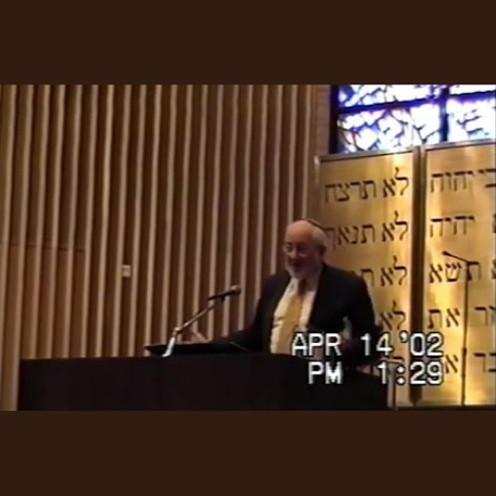 Man speaking at a podium inside a synagogue, with Hebrew text and a stained glass window behind him. Timestamp: April 14, 2002.