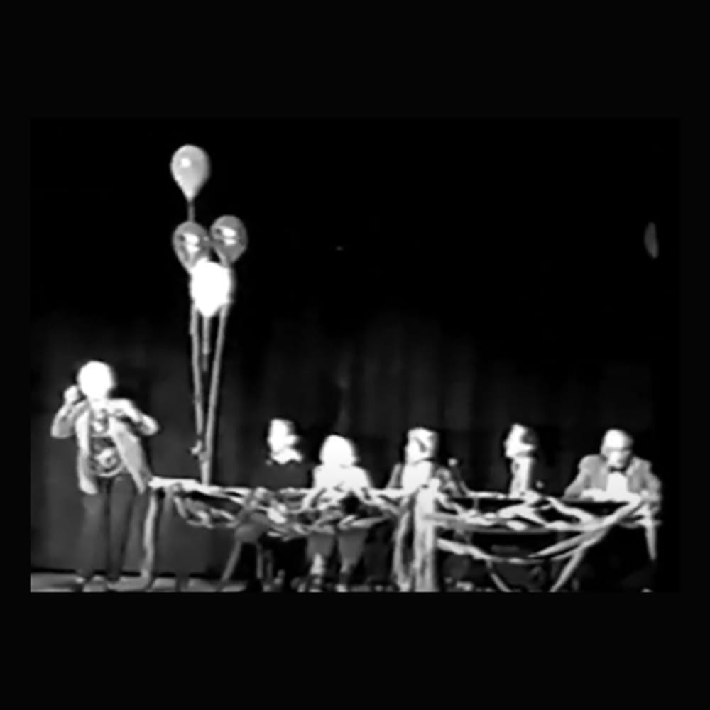 Six people sit at a table onstage with streamers and balloons, while one person stands and speaks into a microphone.