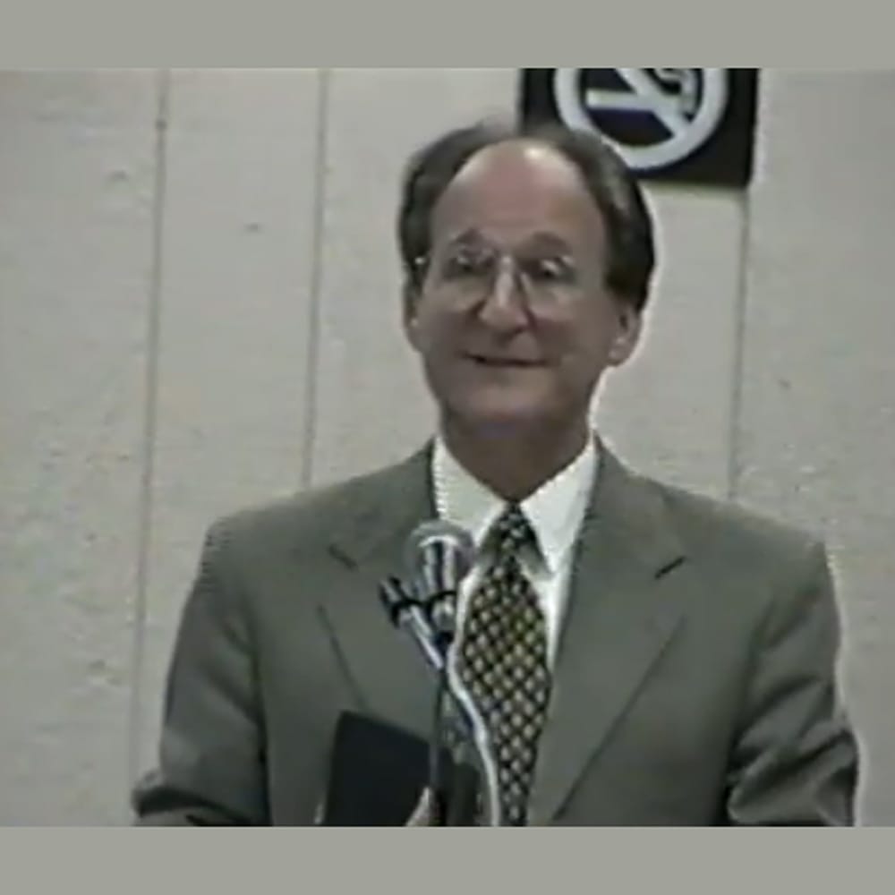 A man in a suit speaks at a microphone indoors, with a no smoking sign visible behind him.