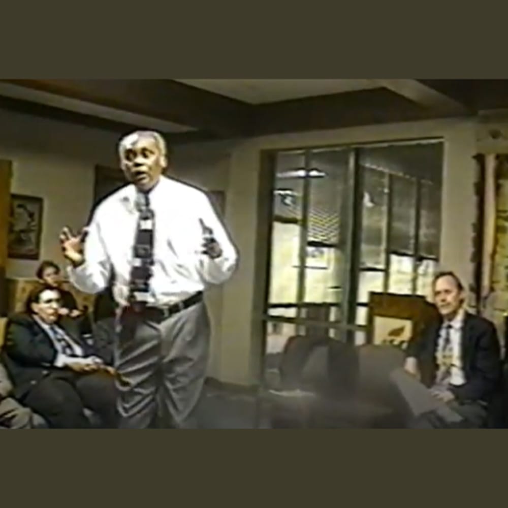 A man in a tie speaks to a seated group in an office or meeting room with glass windows and framed art.
