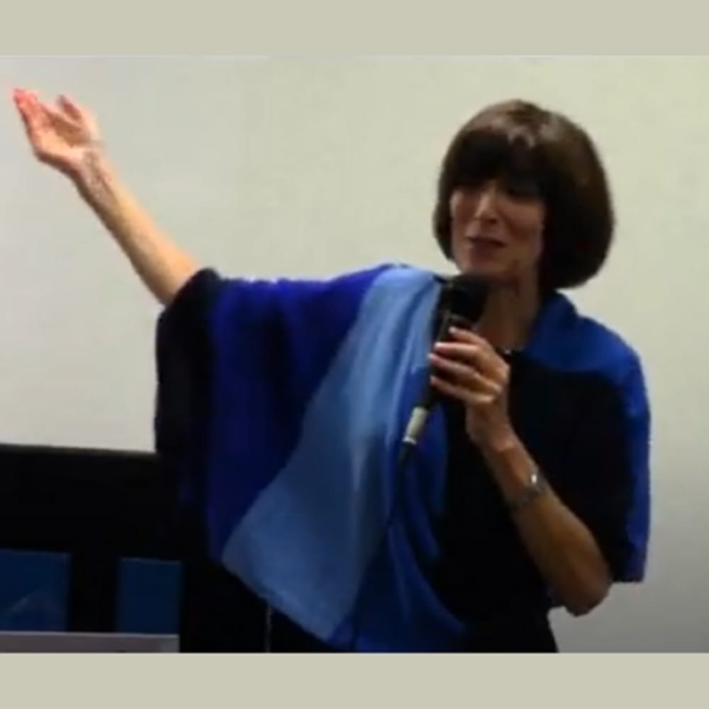 Woman with short dark hair speaks into a microphone, gesturing with her left arm raised and wearing a blue shawl.