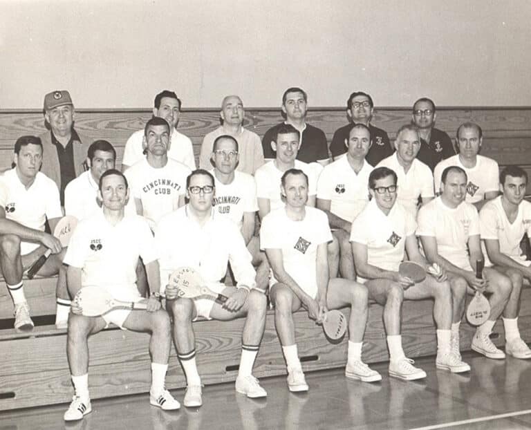 Group of men in sports attire, holding paddles, seated and standing on bleachers in a gymnasium.