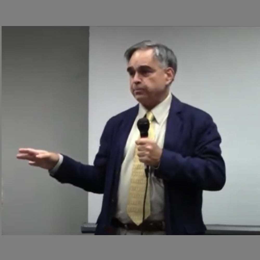 Man in a suit and tie speaking into a microphone, gesturing with his left hand, standing indoors.