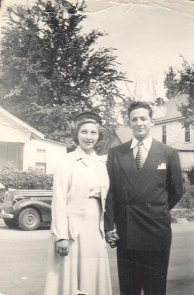 A smiling couple in formal 1940s attire stands holding hands on a suburban street with houses in the background.