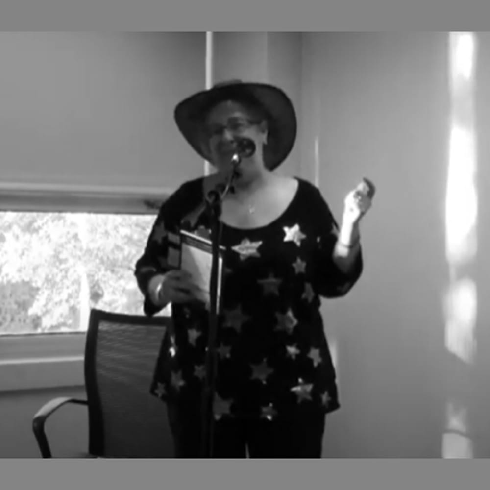 A person in a star-patterned shirt and hat speaks into a microphone while holding a book in a sunlit room.