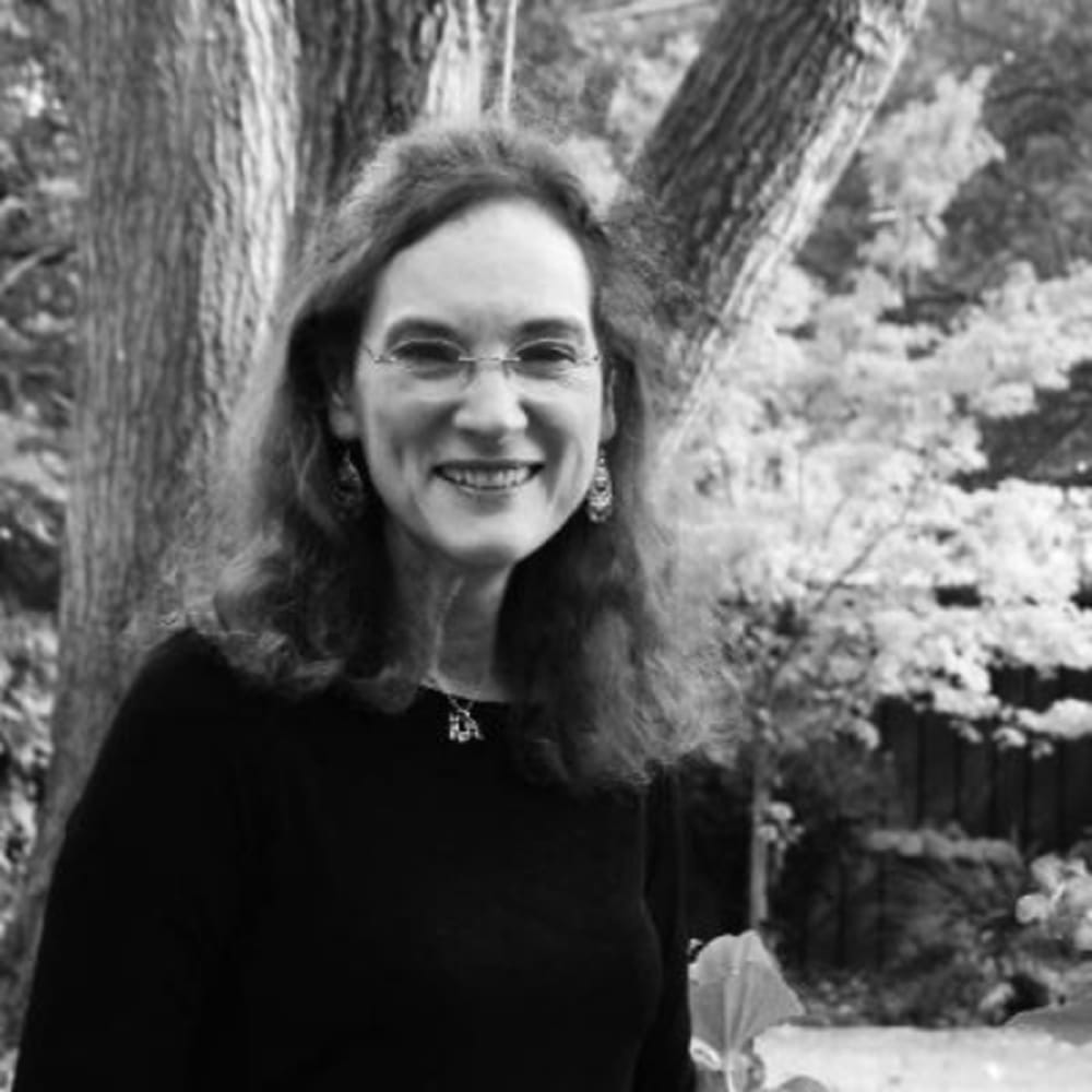 A woman with glasses and curly hair smiles outdoors in front of trees, wearing earrings and a dark top.
