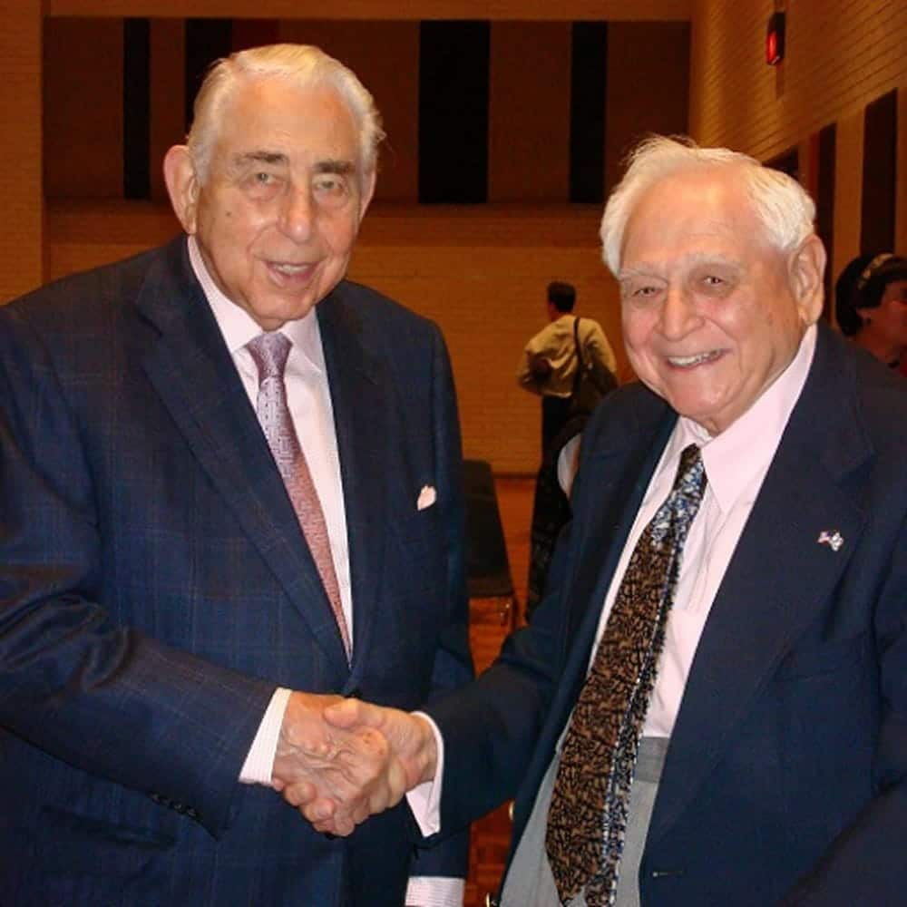 Two elderly men in suits smiling and shaking hands at an indoor event with people in the background.