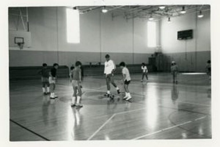 A group of kids play basketball in a gym, with one adult in the center of the court.