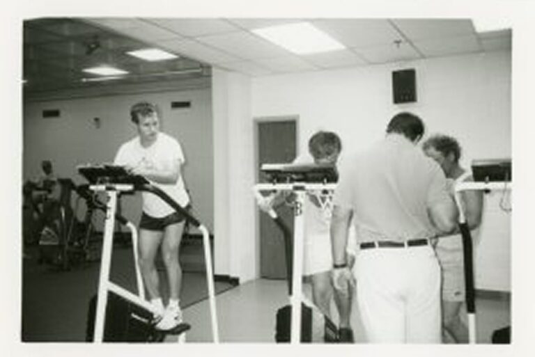 Three people stand on exercise bikes while a man looks at charts in a gym with other people working out in the background.