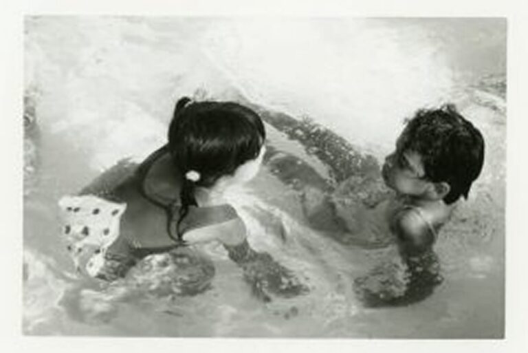 Two young children play and face each other in a swimming pool, water splashing around them.