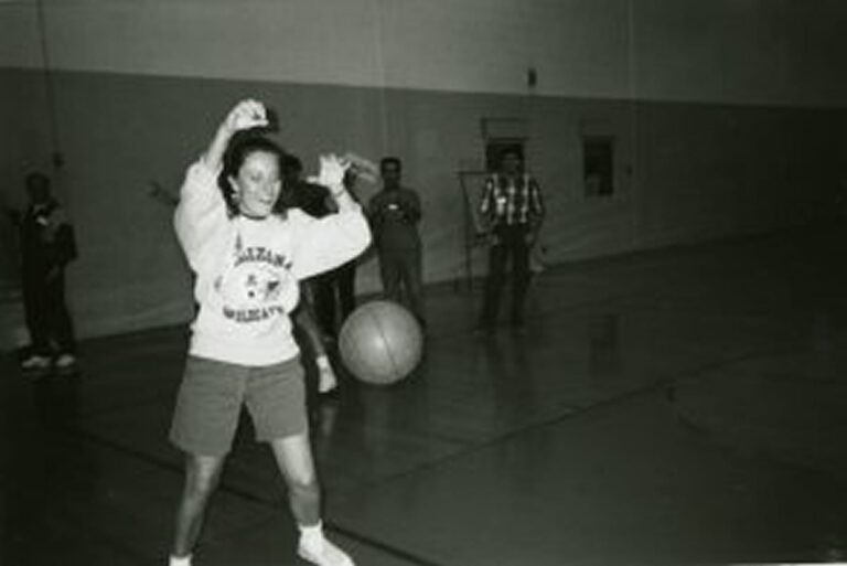 A young woman in a gym prepares to catch a basketball while others watch in the background.