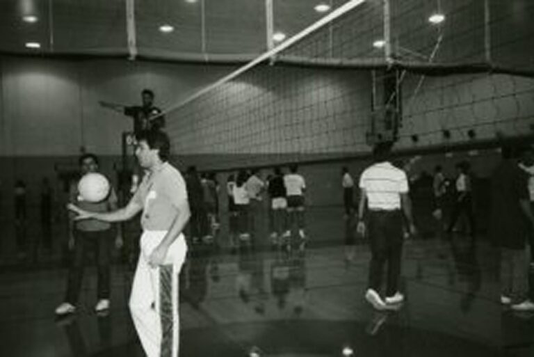 Black-and-white photo of people playing volleyball indoors; one man holds a ball in the foreground.