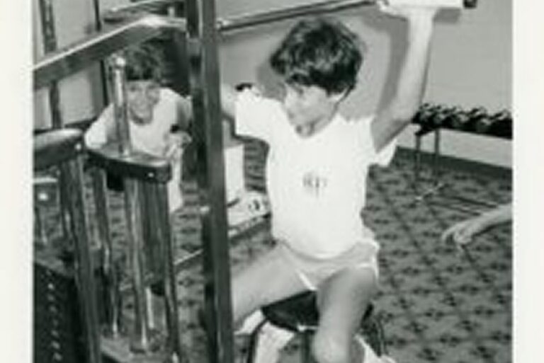 Young boy wearing shorts and a t-shirt uses a weight machine, with his reflection visible in the mirror behind him.