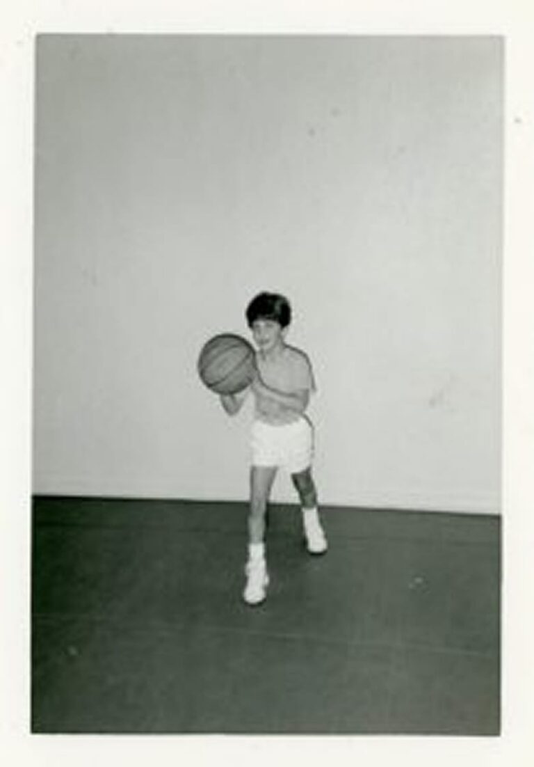 Young child in shorts and sneakers holds a basketball, posed as if about to pass, standing indoors against a plain wall.