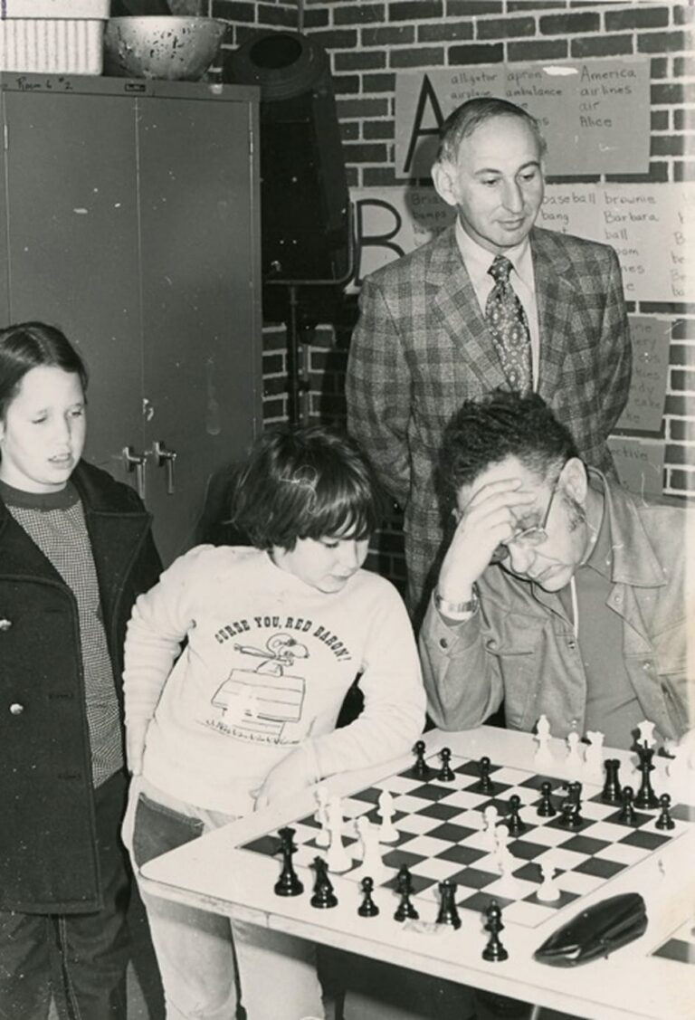 Two children and two adults watch an intense chess game at a table in a classroom with posters on the brick wall.