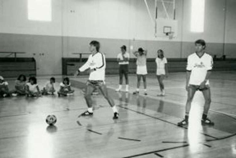 Two teens prepare to kick a soccer ball in a gym while others watch and sit on the floor in the background.