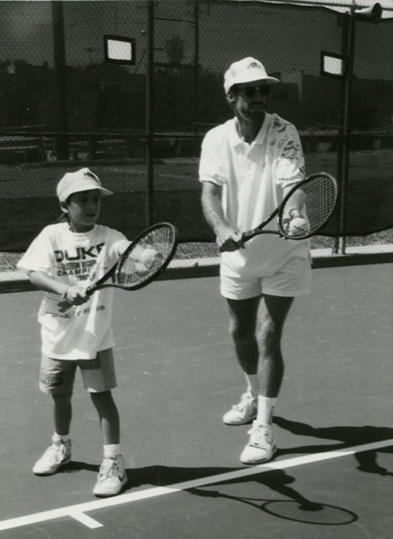 A child and adult play tennis together on an outdoor court, both holding rackets and wearing caps.