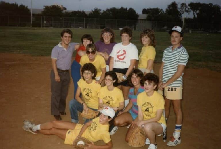 Softball team in yellow shirts poses on a field, with two men standing beside them, all smiling for the photo.