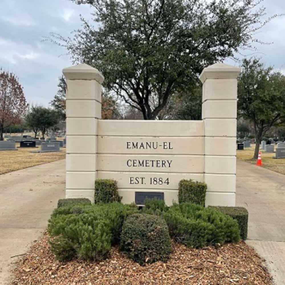 White stone entrance sign reads “Emanu-El Cemetery Est. 1884” with trees and gravestones visible in the background.
