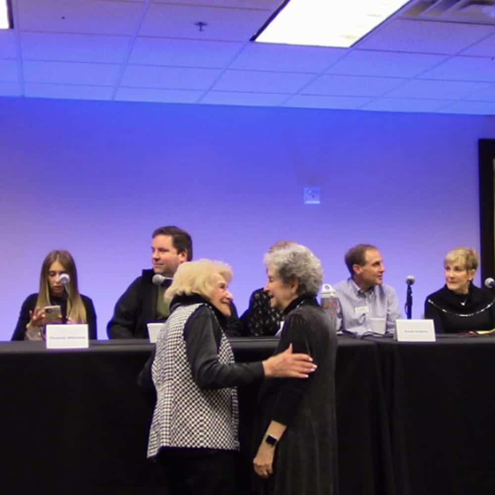 Two women greet in front of a panel of four women and two men sitting at a long table with microphones.