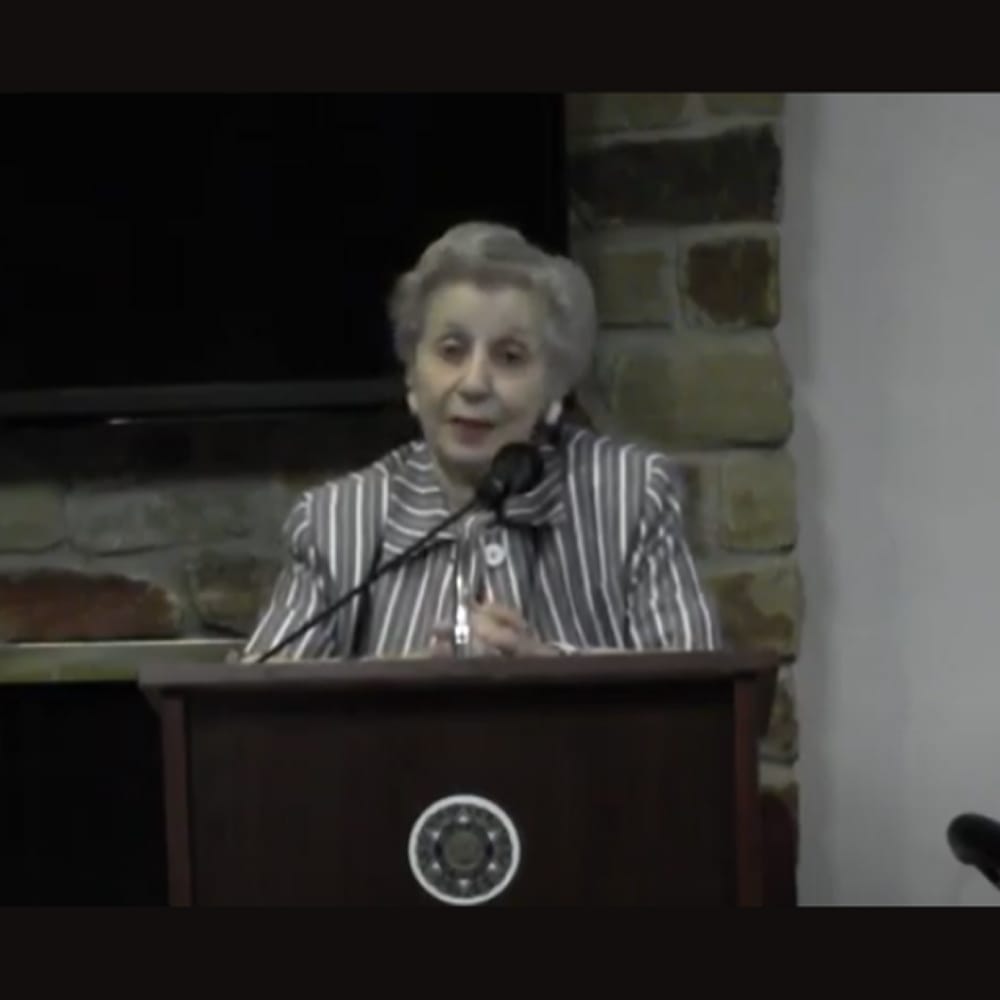 Older woman in a striped jacket speaks at a podium with a microphone, stone wall and TV in the background.