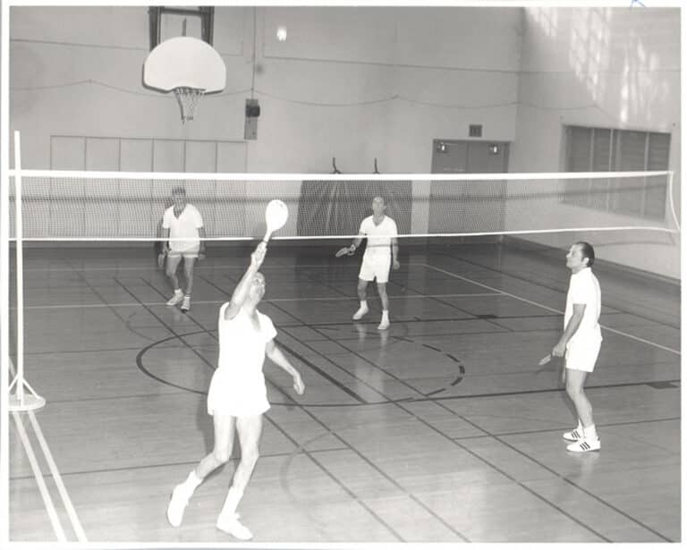 Four men in white athletic gear play badminton in a gymnasium; one is jumping to hit the shuttlecock over the net.