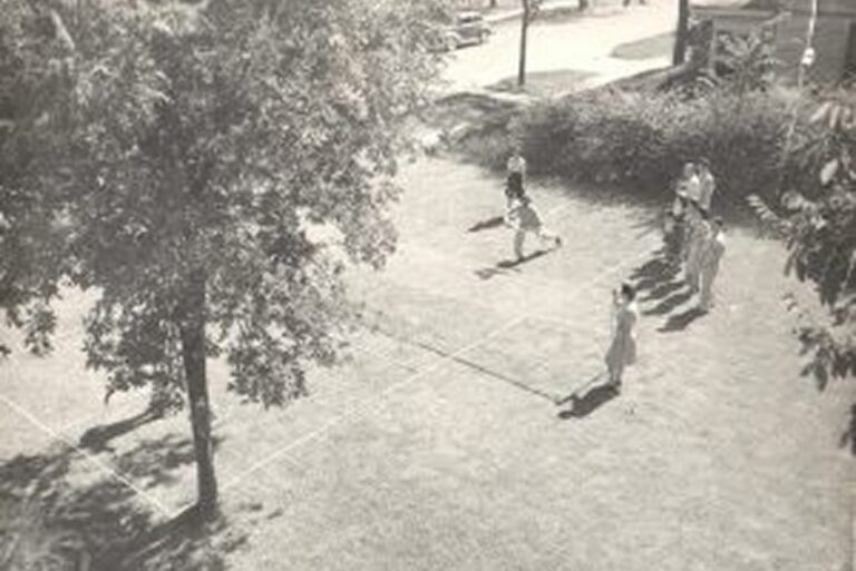 A group of people play badminton on a grassy court under trees, with streets and buildings visible in the background.