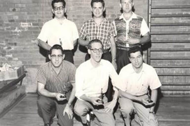 Six men pose indoors, some kneeling and some standing, each holding a small trophy, against a brick wall background.