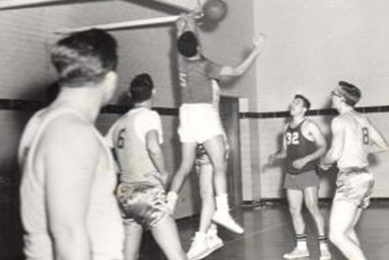 Five men play basketball indoors; one leaps toward the hoop as others watch or try to block him.