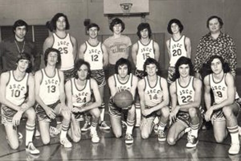 Black and white photo of a vintage basketball team posing in uniform with coaches in a gym.