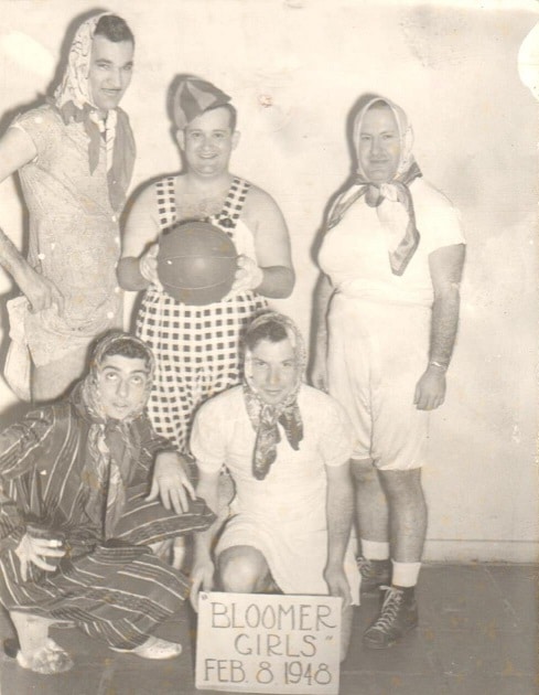 Five men in women’s clothing pose with a basketball; sign reads “BLOOMER GIRLS Feb. 8 1948.”.