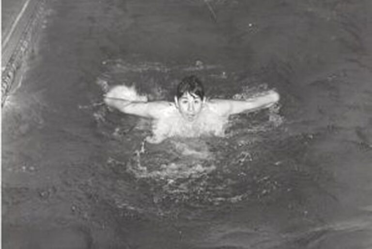 A person swimming the butterfly stroke in a pool, arms extended wide, looking forward, black and white photo.