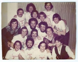 A group of men pose together indoors, many wearing matching white “Christmas Day” t-shirts, smiling at the camera.