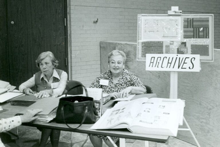 Two women sit at an “Archives” table with open books and a sign in a room with brick walls.